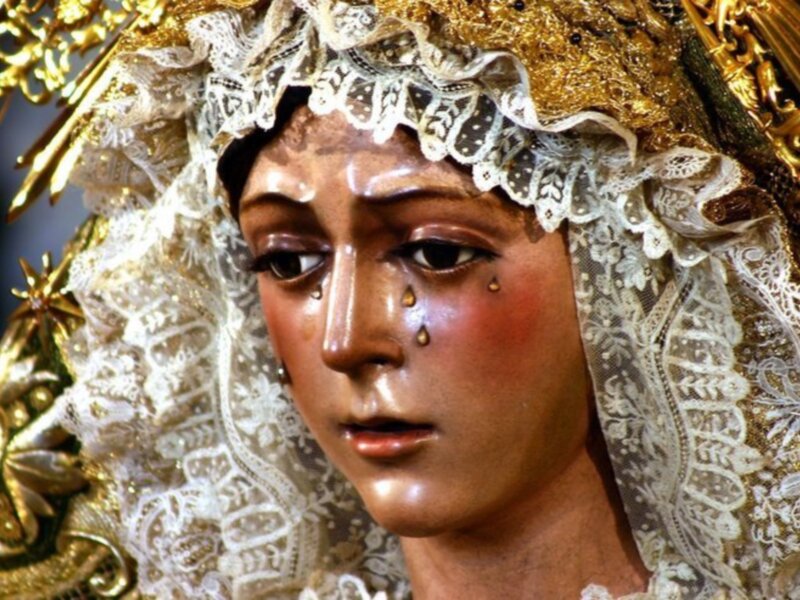 Una figura religiosa con un vestido dorado y una capucha de encaje blanco. La cara está cubierta por un velo que deja solo la parte superior de los ojos y el rostro visible. La expresión parece ser triste o soñolienta, con gotas de lluvia en los párpados. La figura está rodeada por detalles dorados que sugieren un entorno de iglesia o altar.