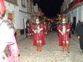Desfile histórico con personajes vestidos como soldados romanos, caminando por una calle adoquinada en la noche.