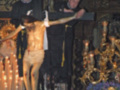 Imagen de un altar con una estatua del Cristo Crucificado, rodeada por velas y adornos dorados. En el fondo, dos personas vestidas formalmente observan la escena religiosa.