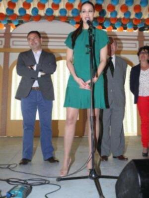 Una mujer en un vestido verde se encuentra frente a un micrófono, mientras otros tres personas observan la escena. La imagen parece ser de una presentación o evento formal en un entorno festivo con adornos colgantes.