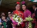 Dos mujeres sonrientes con flores, una vestida de rojo y la otra de verde, posan junto a un niño en un evento festivo.