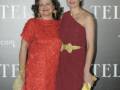Dos mujeres posan en un evento formal, con vestidos rojos y una mano sosteniendo un objeto amarillo.