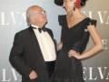 A una mujer y un hombre vestidos formalmente, posando frente a un fondo con el logotipo de Salvatore Ferragamo. La mujer lleva un vestido negro y una sombrero, mientras el hombre está en traje negro con corbata. La escena parece ser de una presentación o evento de moda.