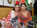 Dos mujeres vestidas con trajes de flamenca tradicionales, sonrientes y sentadas en un ambiente festivo.