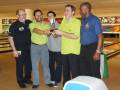 Un grupo de hombres celebrando en un barrio de bowling, uno sostiene un trofeo mientras otros los felicitan.