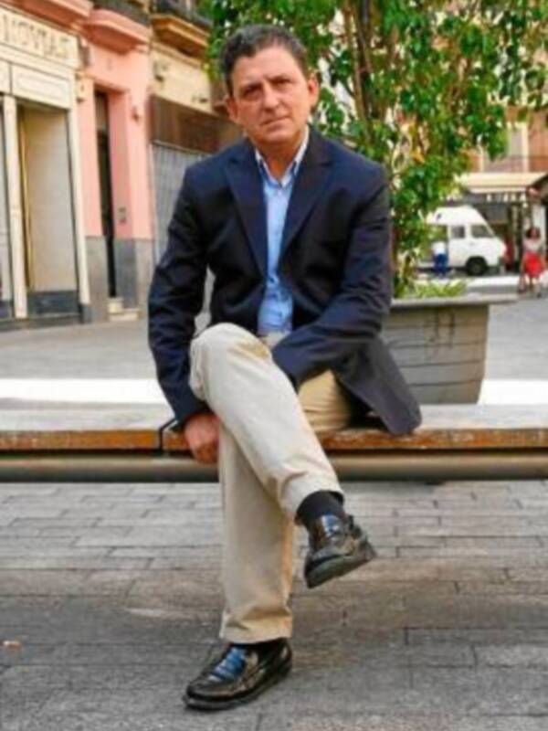 Hombre sentado en un banco de piedra, vistiendo traje y zapatos de cuero, en un entorno urbano con edificios y plantas al fondo.