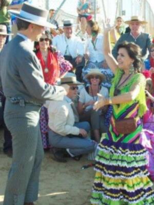 Una mujer en un vestido de colores vibrantes baila en el centro de una reunión festiva, rodeada de personas en trajes tradicionales y sombreros, en un ambiente festivo y colorido.