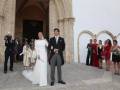 Una boda en un palacio con una pareja recién casada posando frente a una iglesia. La novia lleva un vestido blanco largo y el novio, un traje negro con corbata blanca. A su lado, una cámara toma la foto mientras otras personas observan el evento.