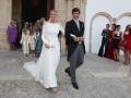 Una mujer vestida de blanco y un hombre vestido formalmente caminan por una calle con piedras. Detrás de ellos, se pueden ver a varias personas vestidas en colores variados, algunas con sombrillas. El entorno parece ser un lugar histórico o ceremonial debido a la arquitectura y el estilo de vestimenta.