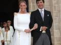 Una pareja en traje y vestido blanco, posando frente a una estructura de piedra.