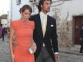 Una pareja elegante camina por una calle adoquinada, ella en un vestido rojo brillante y él en un traje negro con corbata amarilla.