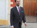 Un hombre vestido con un traje formal camina por una calle con paredes de ladrillo rojo y una puerta metálica.