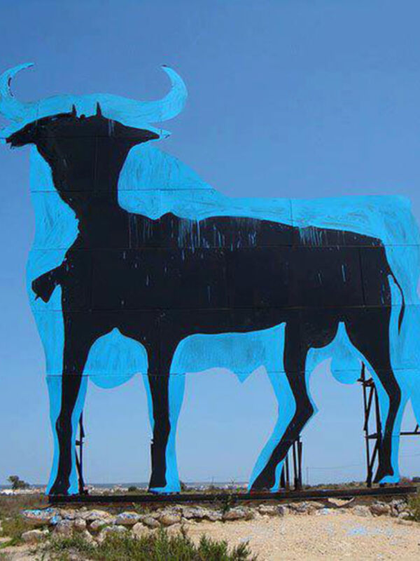 Un gran mural de un toro en una valla de metal, con un cielo azul claro y una casa en el fondo.