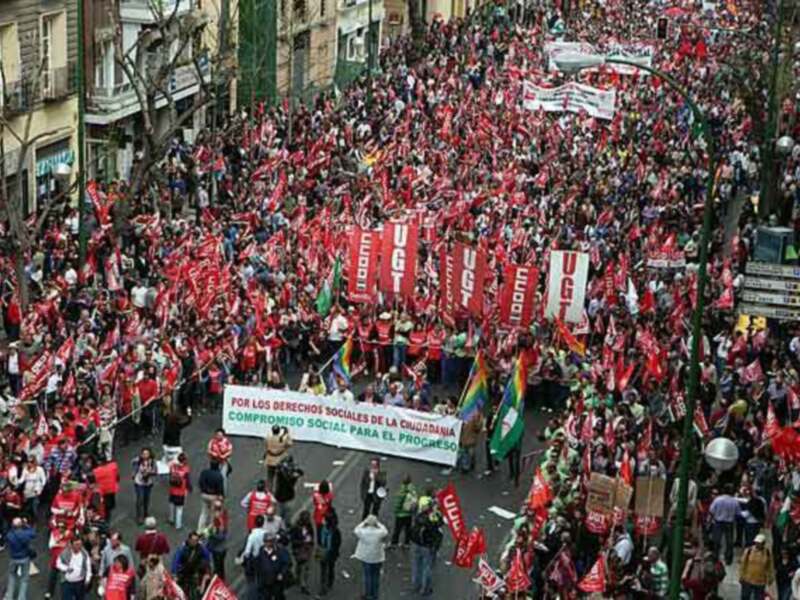Una multitud de personas vestidas con camisetas rojas y pancartas, participando en una manifestación. La imagen muestra a los manifestantes avanzando por una calle con edificios en el fondo, y varios carteles prominentes entre ellos.