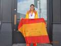 Una joven sostiene una bandera española con mensajes escritos. La imagen muestra un día soleado frente a una puerta de cristal y madera, con el nombre "09.08.2012" en la esquina inferior derecha.