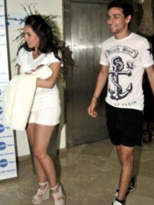 Dos personas caminan juntas, una con un vestido blanco y sandalias, la otra con una camiseta blanca y shorts negros. El fondo muestra un cartel de publicidad.