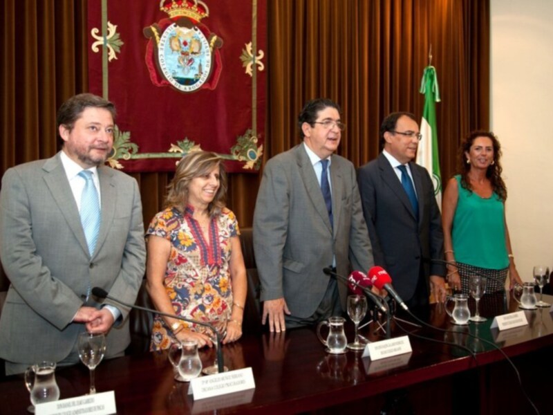Reunión oficial en un salón con escudo y bandera, participantes en trajes formales.