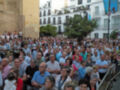 Una multitud de personas reunidas en una plaza, con edificios blancos y azules al fondo. La imagen muestra un ambiente festivo o ceremonial, con gente de todas las edades y estilos.