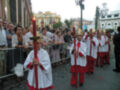 Imagen muestra una procesión religiosa con personas vestidas en trajes rojos y blancos, llevando antorchas. El ambiente es festivo con edificios históricos en el fondo y una multitud observando la celebración.