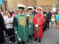 Un grupo de personas vestidas con trajes tradicionales, posiblemente en un evento histórico o ceremonial. La imagen muestra a hombres con vestimentas coloridas y ornamentos, uno de ellos sostiene un bastón. El entorno parece ser una plaza pública con edificios antiguos al fondo y gente observando.