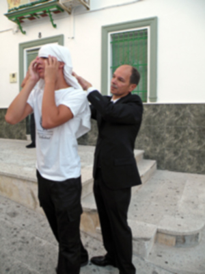 Un hombre en traje negro se acerca a un hombre con una toalla blanca sobre la cabeza, ambos están en una escalera de piedra frente a una casa con ventanas verdes.
