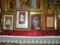 En una mesa cubierta con un mantel rojo y bordado, se colocan cuatro marcos dorados que contienen fotos de personas vestidas con trajes tradicionales, posiblemente en un evento histórico o religioso. En el fondo se pueden ver detalles de una decoración artística y dorada, que añade un ambiente festivo o ceremonial a la escena.