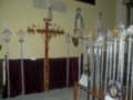 Una exposición de varas procesionales en un espacio decorado con mantones rojos y vitrales. La exposición incluye una cruz de madera en el centro, rodeada por varias varas procesionales ornamentadas con detalles metálicos y cristales. En el fondo, se pueden ver vitrales representando a personajes religiosos y una pintura mural.