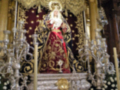 Una estatua religiosa en un altar dorado, rodeada de candelabros y flores. La estatua está vestida con un manto rojo y una corona, con detalles dorados que reflejan la solemnidad del lugar. El altar está decorado con candelabros de cristal y flores blancas, creando un ambiente de reverencia.