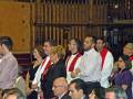 Una imagen de una ceremonia religiosa donde una persona con un velo rojo se encuentra entre otros individuos vestidos formalmente. La escena parece ser en un lugar de culto, posiblemente una iglesia, con estructuras de madera y luces de techo.