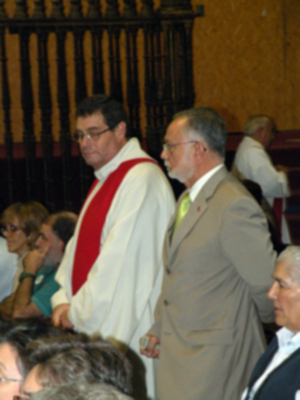 Persona vestida con un hábito rojo y blanco, posiblemente un sacerdote, caminando en una ceremonia o evento formal en un interior con asistentes.