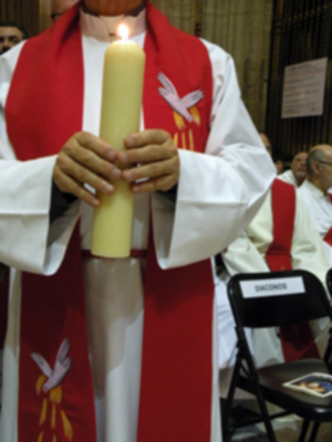 Un sacerdote sostiene una vela amarilla en un entorno religioso, con vestimenta sacerdotal y una saya roja.