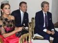 Una mujer con un vestido de encaje negro y una corona, junto a dos hombres en trajes formales sentados en sillas doradas.