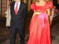 Una mujer viste un vestido rojo con detalles amarillos y una corbata rosa. Un hombre en traje negro la acompaña. La escena parece ser un evento formal al aire libre con una estructura de madera y vidrio en el fondo.
