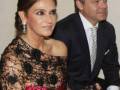 Una mujer con un vestido negro y rosa, lleva una corona de flores en su cabeza. Un hombre junto a ella, con traje negro y corbata blanca. Ambos están en un evento formal.
