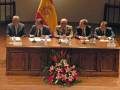 Una reunión formal en un salón con una bandera española, cuatro hombres sentados a una mesa de madera, con un centro floral en el frente.