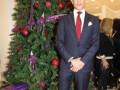 Elegante hombre en traje de negocios posando frente a un árbol de Navidad decorado con bolas rojas y regalos, en un ambiente festivo y formal.