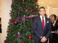 Elegante hombre en traje de negocios posando frente a un árbol de Navidad adornado con bolas rojas y purpuras.