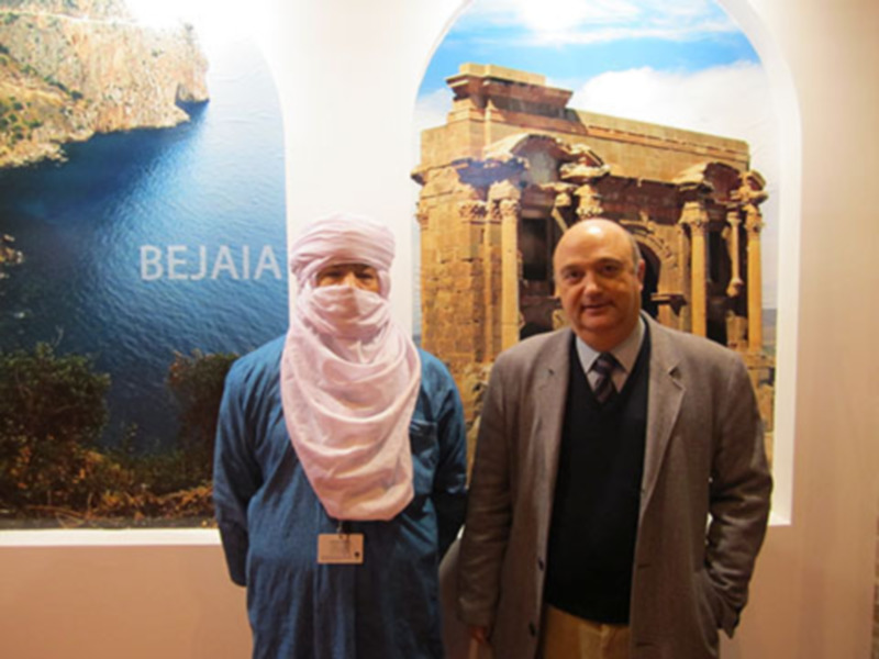 Dos personas posan frente a un cartel con imágenes de la costa y ruinas de Bejaia, Argelia.