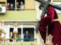 Una imagen de una procesión religiosa, con una figura vestida de rojo cargando una cruz, mientras otros observan desde una ventana.