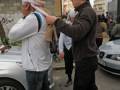 Un hombre con una camiseta blanca y un pantalón negro sostiene un micrófono, mientras otro hombre con una camiseta marrón y pantalones negros lo mira. En el fondo, se ven dos coches estacionados y una pared con grafitis.
