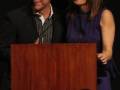 Una mujer sonríe mientras se encuentra junto a un hombre en una presentación formal. Ambos están detrás de un podio marrón, con el hombre vestido en una camisa a rayas y la mujer en un vestido azul. La imagen sugiere una celebración o evento formal, posiblemente un congreso o conferencia.