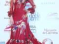 Una mujer con un vestido rojo flamenco largo y elaborado, adornado con flores y detalles dorados. La imagen está en un escenario con logotipos de marcas y texto que indica el evento "Salón Internacional de la Moda Flamenca 2013".
