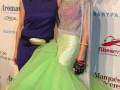 Dos mujeres posan en un evento, una con vestido azul y la otra con vestido verde y rosa. Detrás de ellas, se puede ver un cartel que dice "SIMOF 2013" y "Marques de Cáceres".
