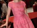 A una mujer con un vestido rosa claro que tiene detalles de encaje. Está posando frente a una pared roja y parece estar en un evento formal.