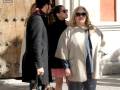 Dos personas caminan por una calle, una de ellas con un abrigo beige y una falda rosa, mientras la otra lleva un abrigo oscuro y una blusa negra.