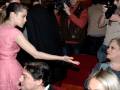 Una mujer en un vestido rosa se acerca a una persona sentada, mientras otros fotógrafos la filman. La escena parece ser en un evento formal o de premiación.