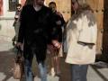 Dos personas caminan por una calle adoquinada, intercambiando una conversación amistosa. El hombre lleva un abrigo negro y elige, mientras la mujer, con un abrigo beige, observa. En el fondo, se pueden ver otras personas y una puerta de madera tallada.