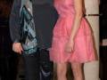 Dos personas posan juntas en una alfombra roja, uno con un vestido de lentejuelas y pantalones negros, el otro con un vestido rosa. Detalle de la alfombra roja y una columna en el fondo.