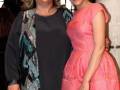 Dos mujeres posando juntas en un evento, una con vestido rosa y otra con gafas de sol y chaleco bordado.
