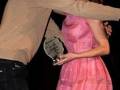 Un hombre abraza a una mujer en un evento, ella sostiene un premio.